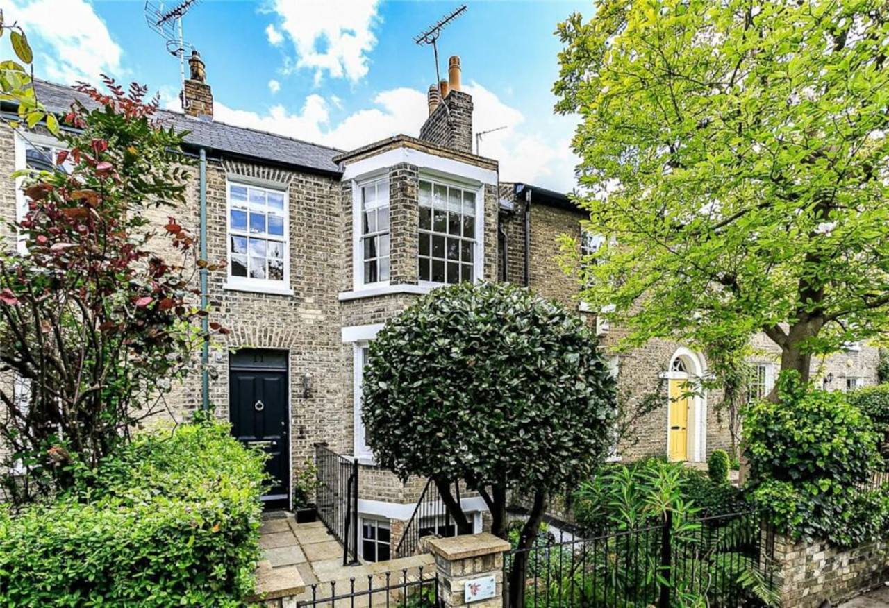 Exterior photograph of a traditional brick terraced house in Cambridge with a black front door, bay window, and front garden with mature greenery