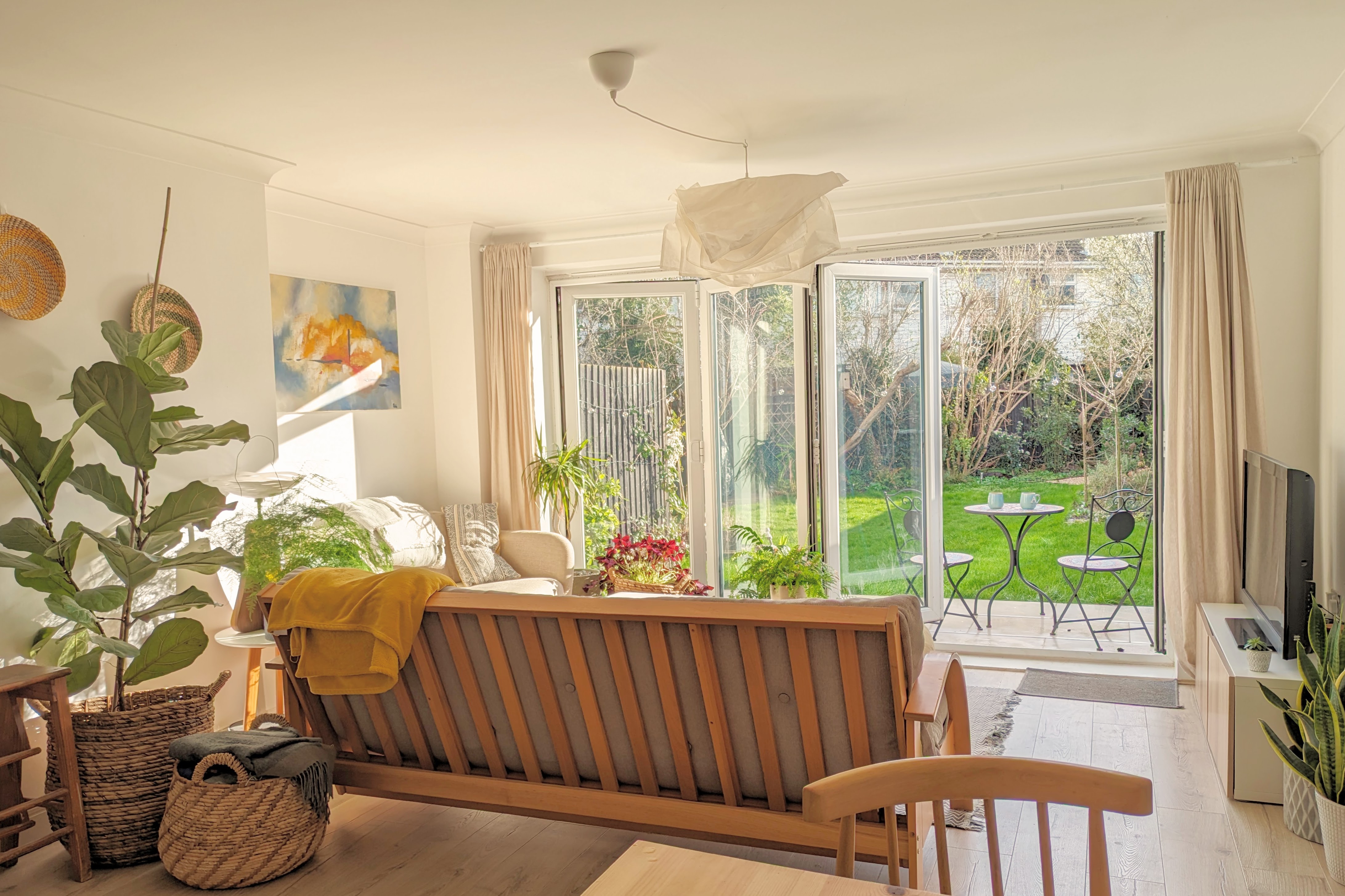 A bright, sunlit living room featuring a wooden sofa, indoor plants in woven baskets, and large folding glass doors opening onto a green garden patio
