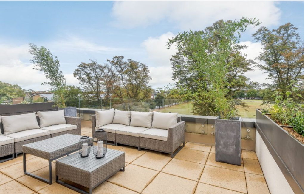 A professional photograph of a spacious rooftop terrace featuring grey wicker outdoor sofas with beige cushions, matching coffee tables with candle lanterns, and an urban view